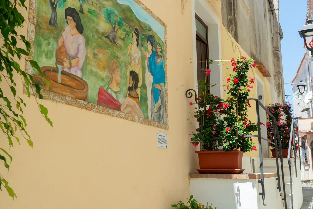 Callejuela del pueblo con mural, flores rojas en maceta y barandilla de hierro