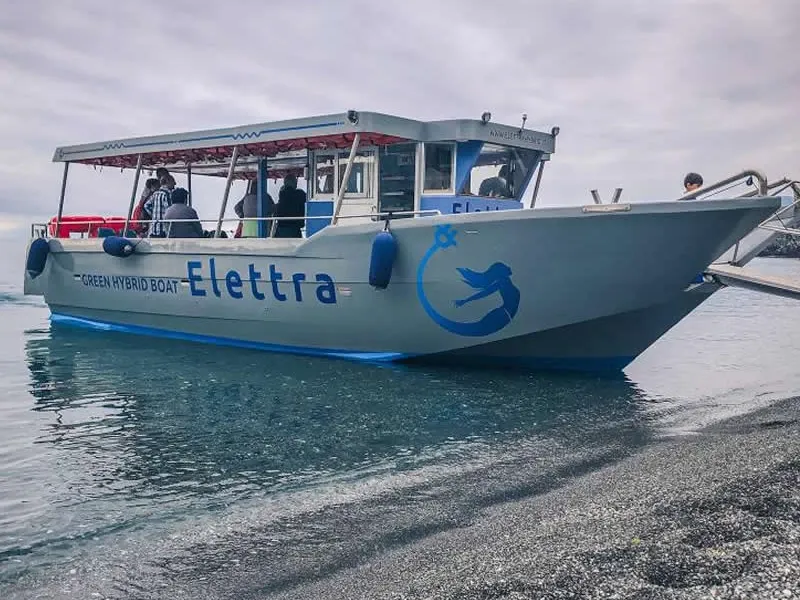 La barca 'Elettra' con pasajeros junto a una playa de guijarros oscuros
