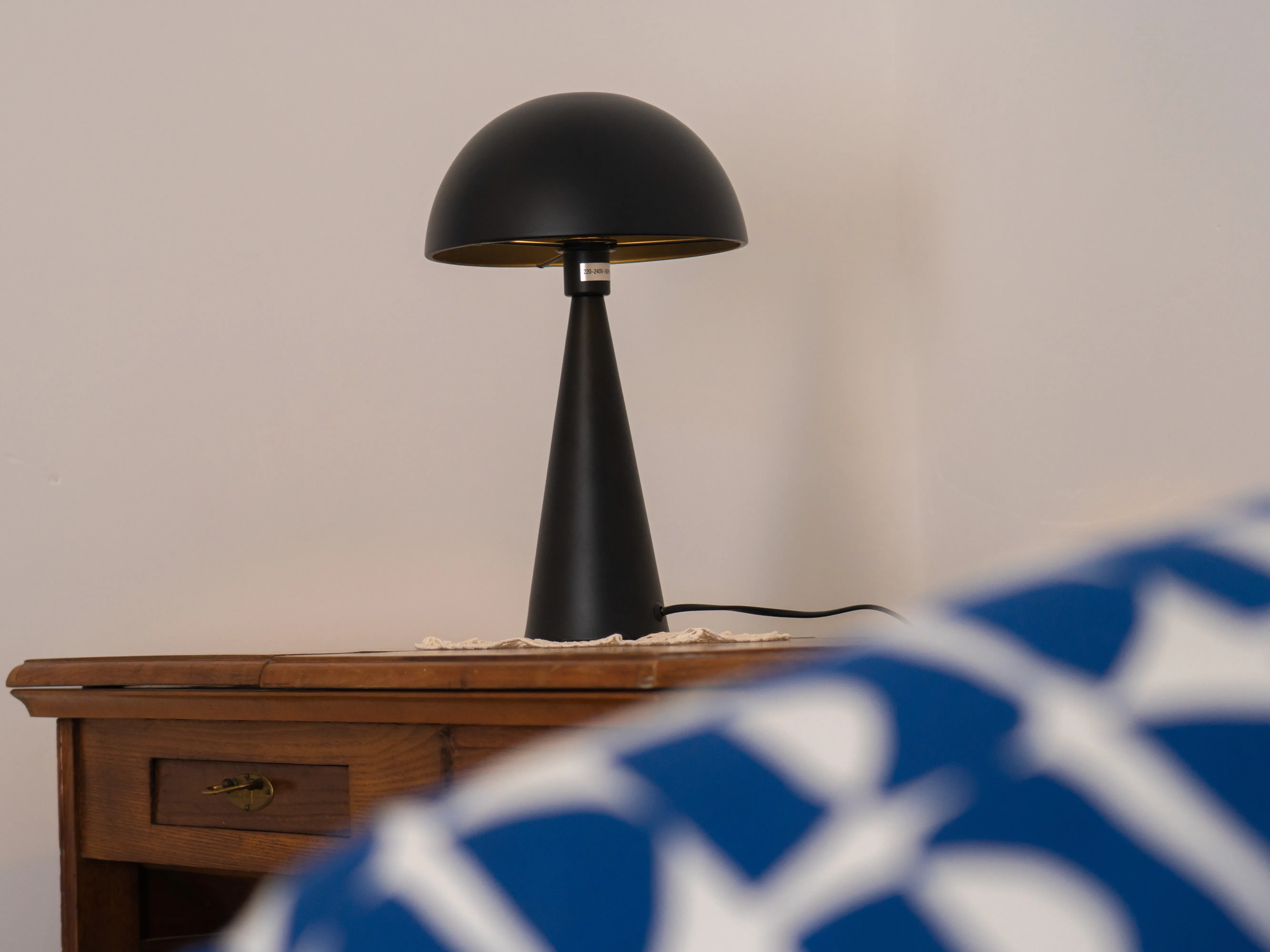 Lámpara negra tipo seta sobre una mesilla de madera, delante un cojín azul y blanco