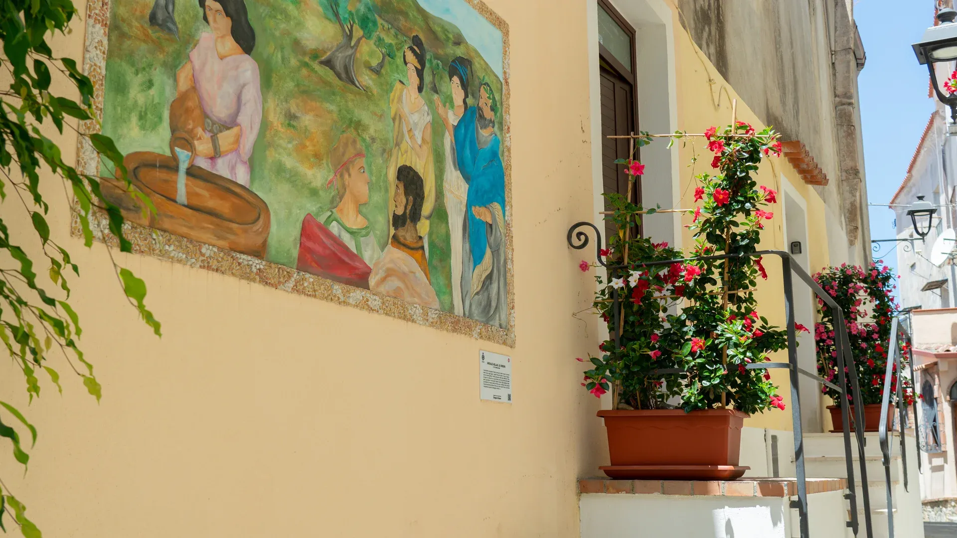 Fachada amarilla con mural y placa, dos macetas de flores rojas y escalones blancos