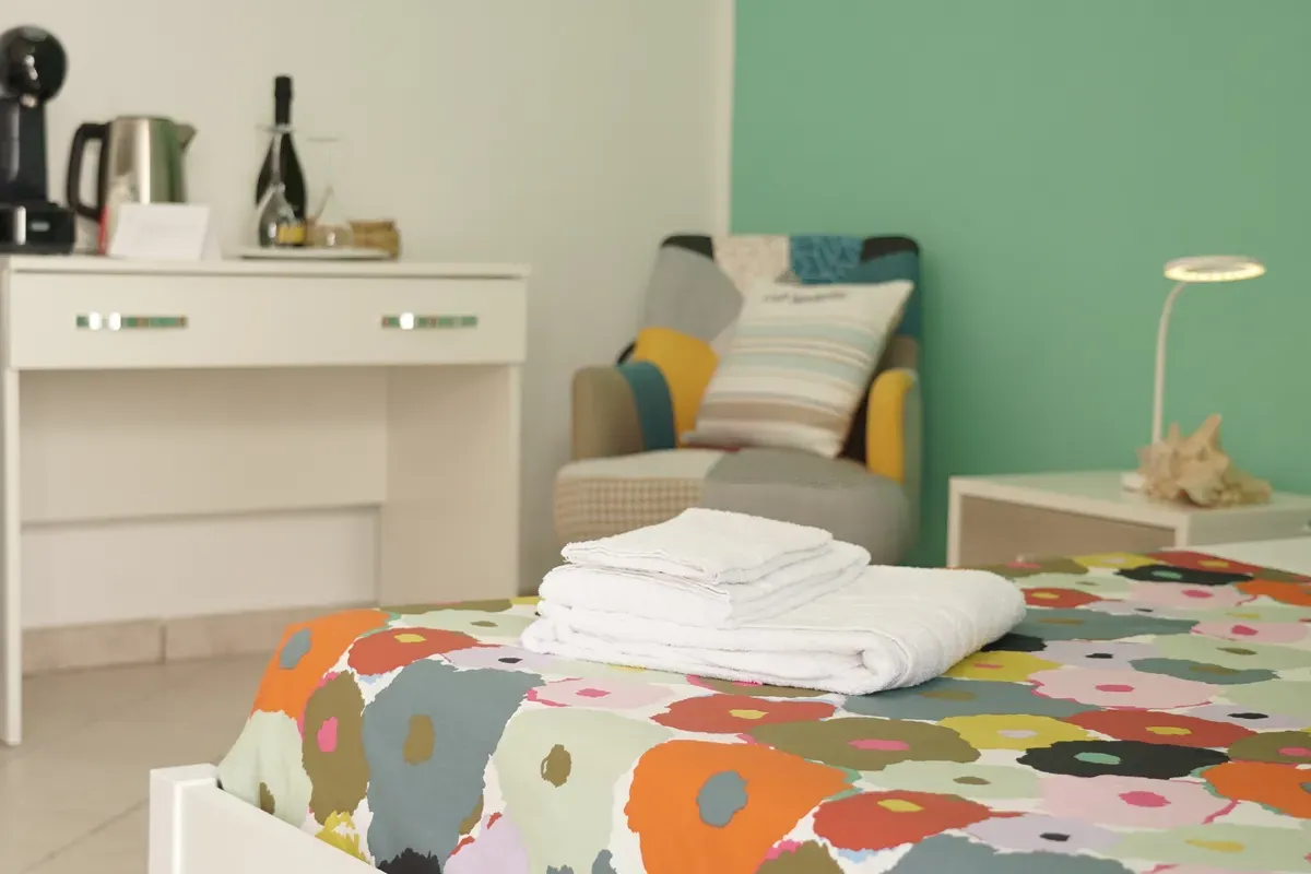 Cama con colcha de flores y toallas, sillón de patchwork y escritorio con hervidor